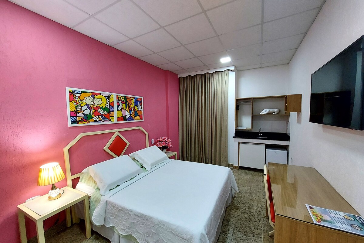 A hotel room with a bed, bedside table, lamp, artwork, curtains, a desk, a mirror, and a TV.