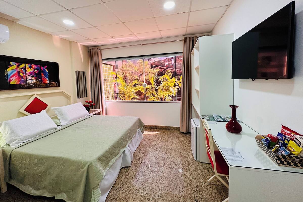 Habitación de hotel con cama, escritorio, televisión, ventana con vista a árboles y un cuadro en la pared.