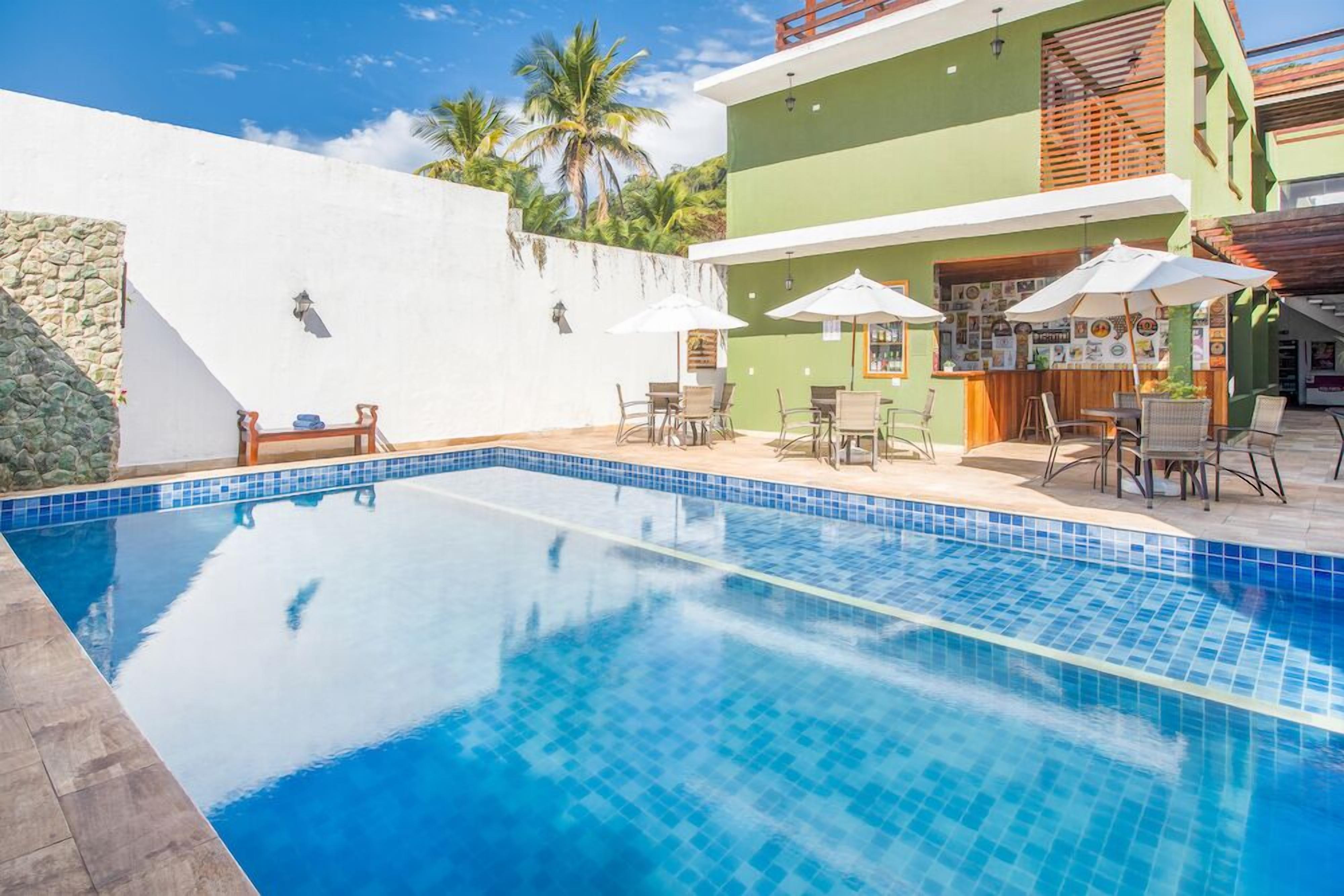 Una piscina con bordes azulejados, rodeada de mobiliario exterior y sombrillas, con un edificio al fondo.