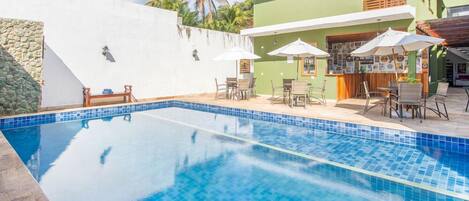 Una piscina con bordes azulejados, rodeada de mobiliario exterior y sombrillas, con un edificio al fondo.