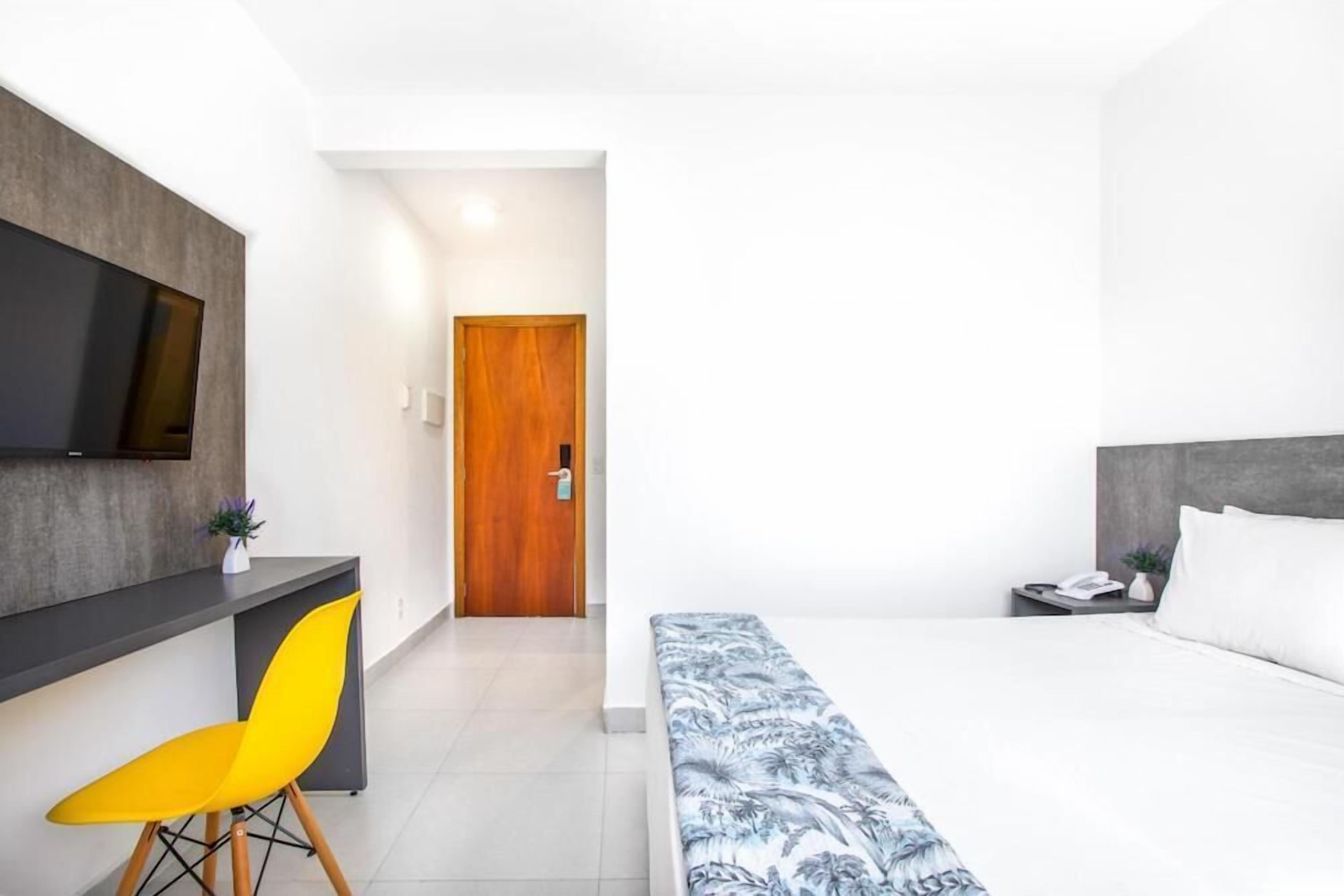 Habitación de hotel moderna con una cama, un escritorio con una silla, un televisor montado en la pared y una puerta de madera.
