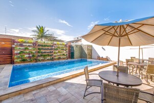 Una piscina con borde de azulejos, rodeada de muebles de mimbre y un paraguas grande.