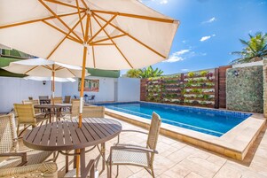 Zona de piscina con mesa y sillas de madera, un gran paraguas y una pared decorada con plantas.