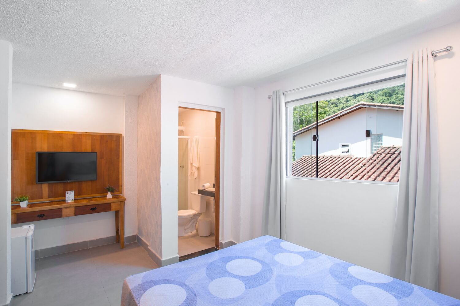 Un dormitorio moderno con televisor, un escritorio de madera y un baño visible a través de una puerta abierta.