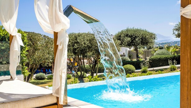 Ein Pool mit Wasserspiel, umgeben von viel Grün und blauem Himmel.