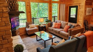 Ein gemütliches Blockhaus-Interieur mit Kamin, großen Fenstern, einer Couch und einem Fernseher.