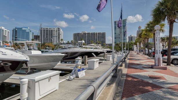 A marina with several boats docked, a paved walkway, and a city skyline in the background. - Spacious 2 bedroom+ den condo in Arielle on Palmer Ranch condominiums for rent (Sarasota)