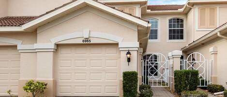 A beige two-story house with a garage number 6965, a gated entrance, and a paved driveway.