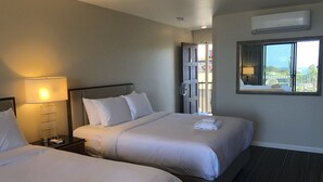 A hotel room with two beds, a nightstand with a lamp, an air conditioning unit, and a window with a view. - Moonlight Beach Motel (Encinitas)