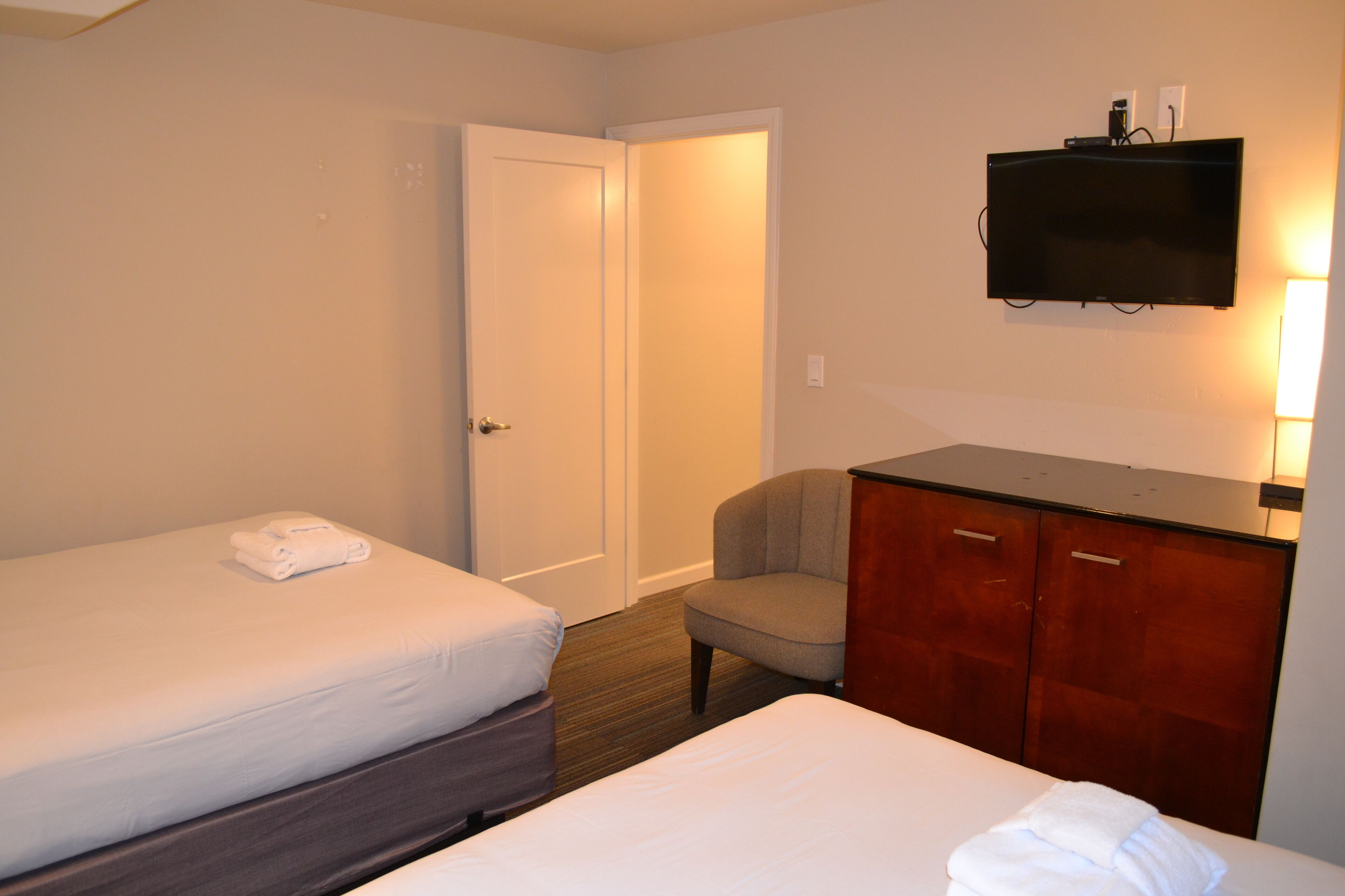 A hotel room with two beds, a chair, a desk with a flat-screen TV, and a wardrobe.