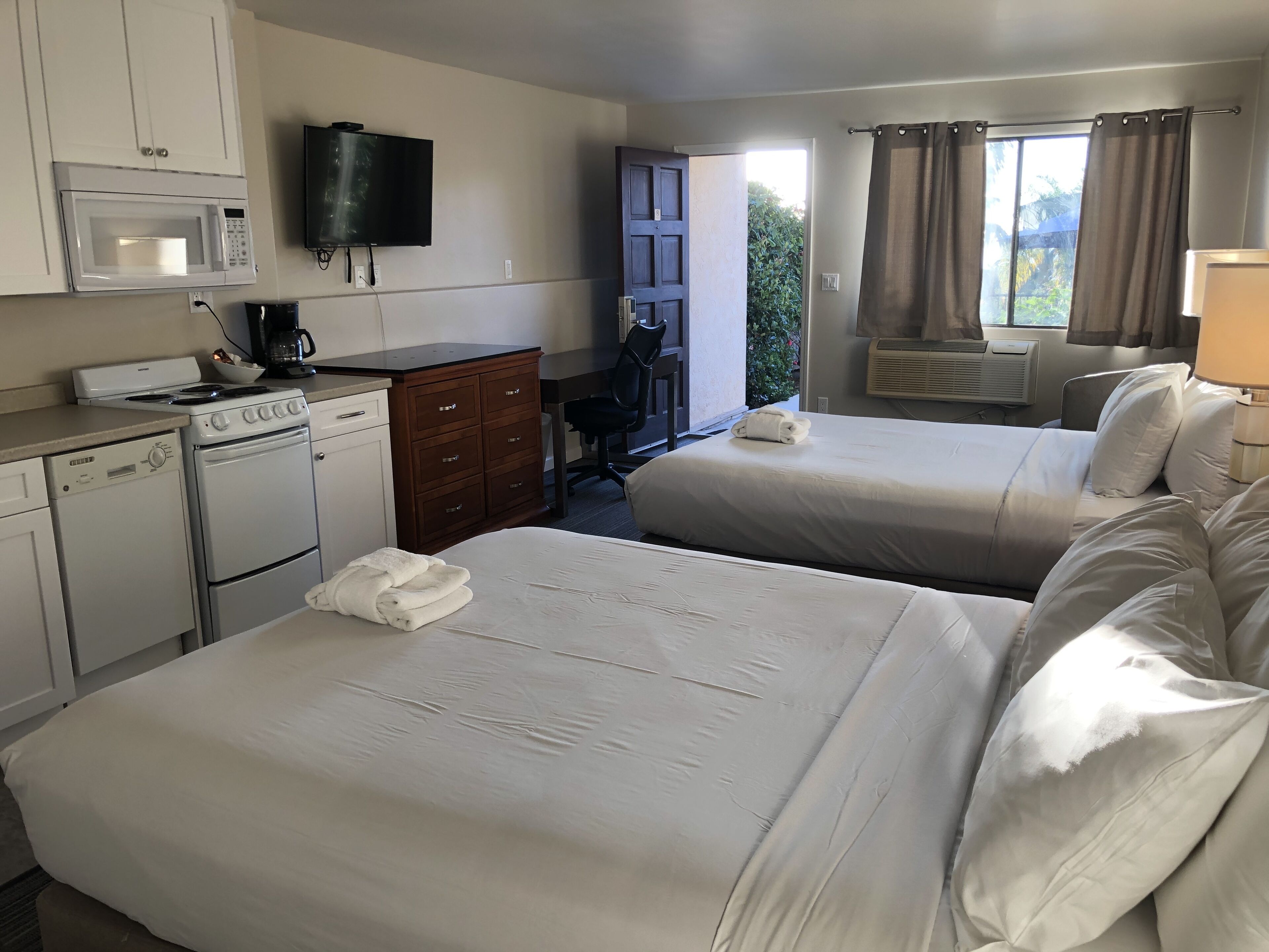 Hotelkamer met twee bedden, een kitchenette met magnetron, kookplaat en koffiezetapparaat, een televisie en een raam met gordijnen.