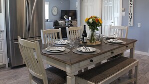 Ein Esszimmer mit einem Holztisch, gedeckt für vier Personen, umgeben von weißen Stühlen. Auf dem Tisch steht eine Vase mit Blumen, und es befindet sich eine Weinflasche. Die Wände sind hellblau und die Decke weiß.