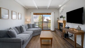 A living room with a grey sofa, a wooden coffee table, a TV, and a balcony with a view of trees and mountains.