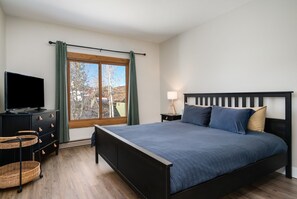 Ein Schlafzimmer mit Bett, Fernseher, Schminktisch, Lampe und einem Fenster mit Vorhängen.