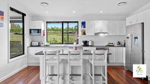 A modern kitchen with white cabinetry, a large island with bar stools, a built-in oven, and a view of a vineyard through the window. - iyla Estate - Your exclusive Vineyard & Cottage stay @ Pokolbin, Hunter Valley! (Pokolbin)