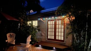Une terrasse en bois avec deux chaises blanches et un banc, une maison en pierre avec une porte en verre, et des guirlandes lumineuses au-dessus de la porte.
