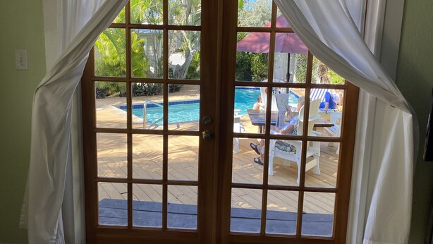 Une porte en bois, ornée de rideaux blancs, donne sur un espace piscine avec une terrasse en bois, des chaises longues et un parasol rouge.