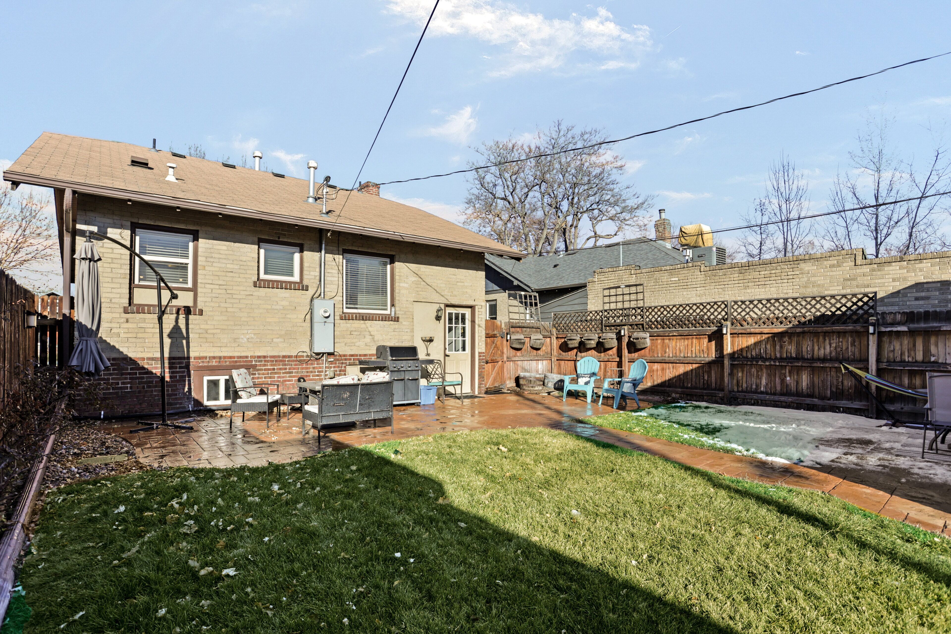 A residential backyard with a brick house, a patio area, a barbecue grill, and a wooden fence.