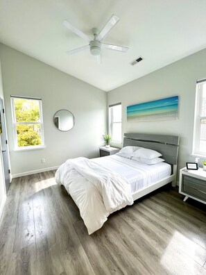 Un dormitorio con cama, mesita de noche, ventilador de techo, ventana y un cuadro.