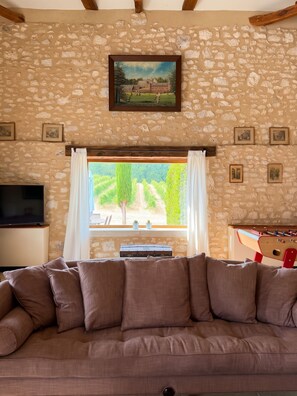A living room with a stone wall, a sofa, a TV, a window with curtains, and a painting above the window. - Homerez - "Sigoulès villa with pool" (Sigoulès)
