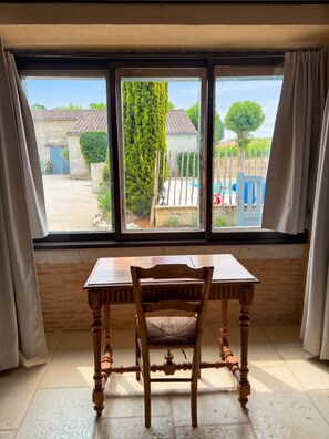 A room with a wooden table and chair, a window with curtains, and a view of a garden and pool.