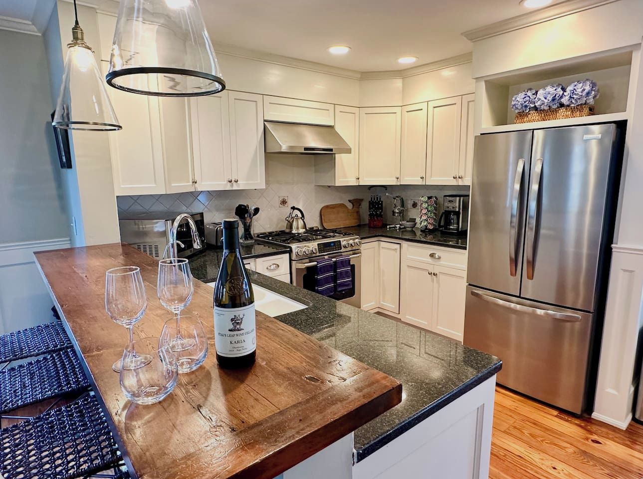 A modern kitchen with white cabinets, a stainless steel refrigerator, and a wooden island with wine glasses and a bottle.