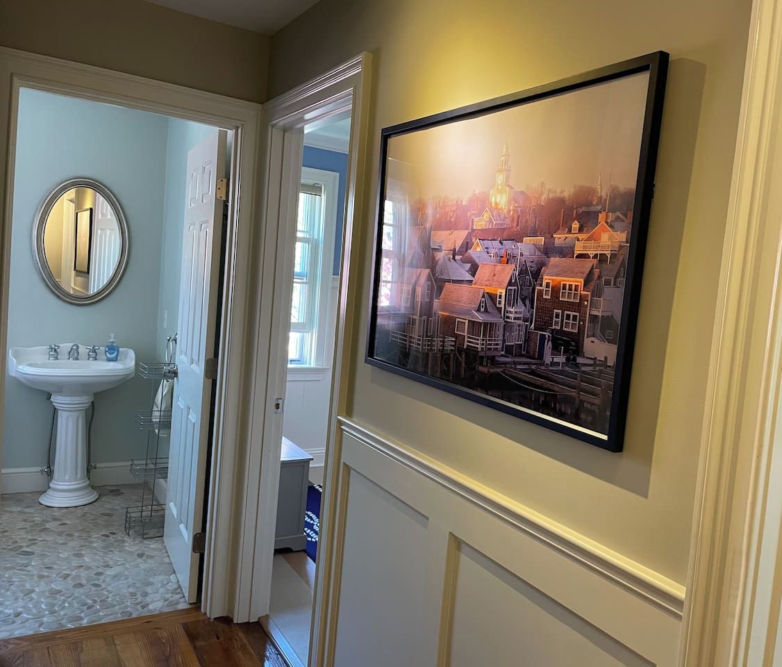 Un corridoio con un lavabo a piedistallo bianco, uno specchio rotondo e un quadro incorniciato con una vista cittadina sulla parete.