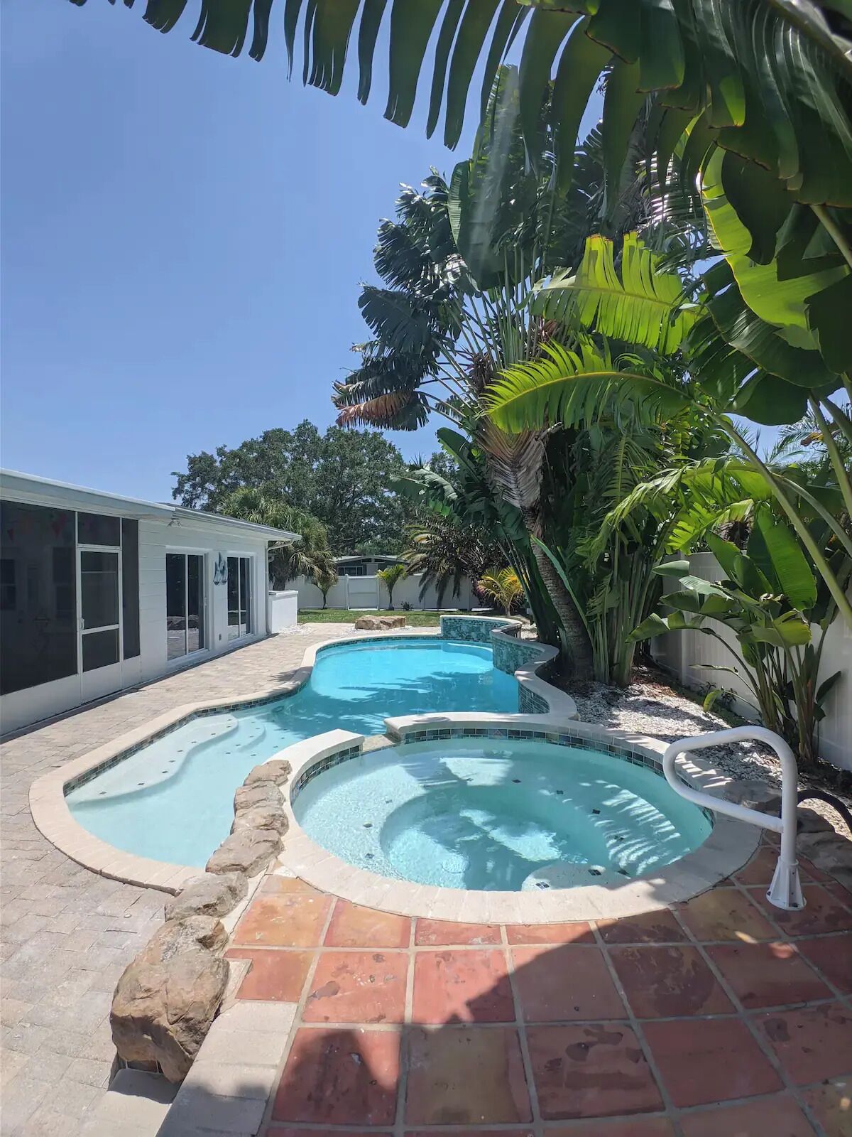 Uma piscina com design curvo, rodeada por azulejos de pedra e plantas tropicais.