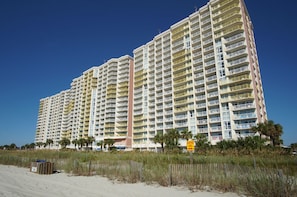 A high-rise hotel with multiple balconies, a sandy beach in the foreground, and a 'Surfing Area' sign.