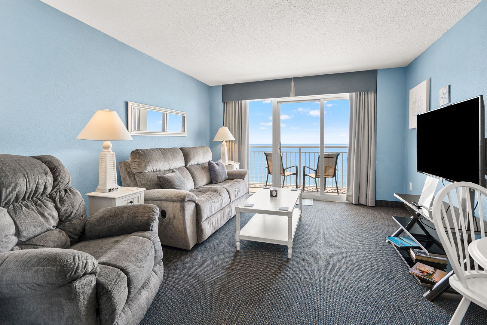 A hotel room with a sofa, armchair, coffee table, TV, and a balcony with a view.