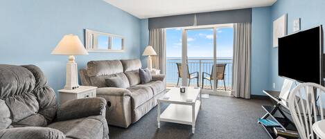 A hotel room with a sofa, armchair, coffee table, TV, and a balcony with a view.