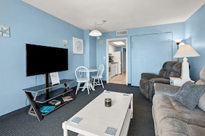 A hotel room with a flat-screen TV, a white coffee table, a sofa, a dining area with white chairs, and a lamp.