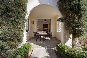 Une terrasse extérieure avec une table et des chaises, entourée de verdure et dotée d’une porte en arc donnant sur une pièce.
