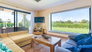 A modern living room with a sofa, a coffee table, and a TV. There is a large window with a view of greenery. - YourHouse Villa Romani con piscina privada y jardín (Muro)