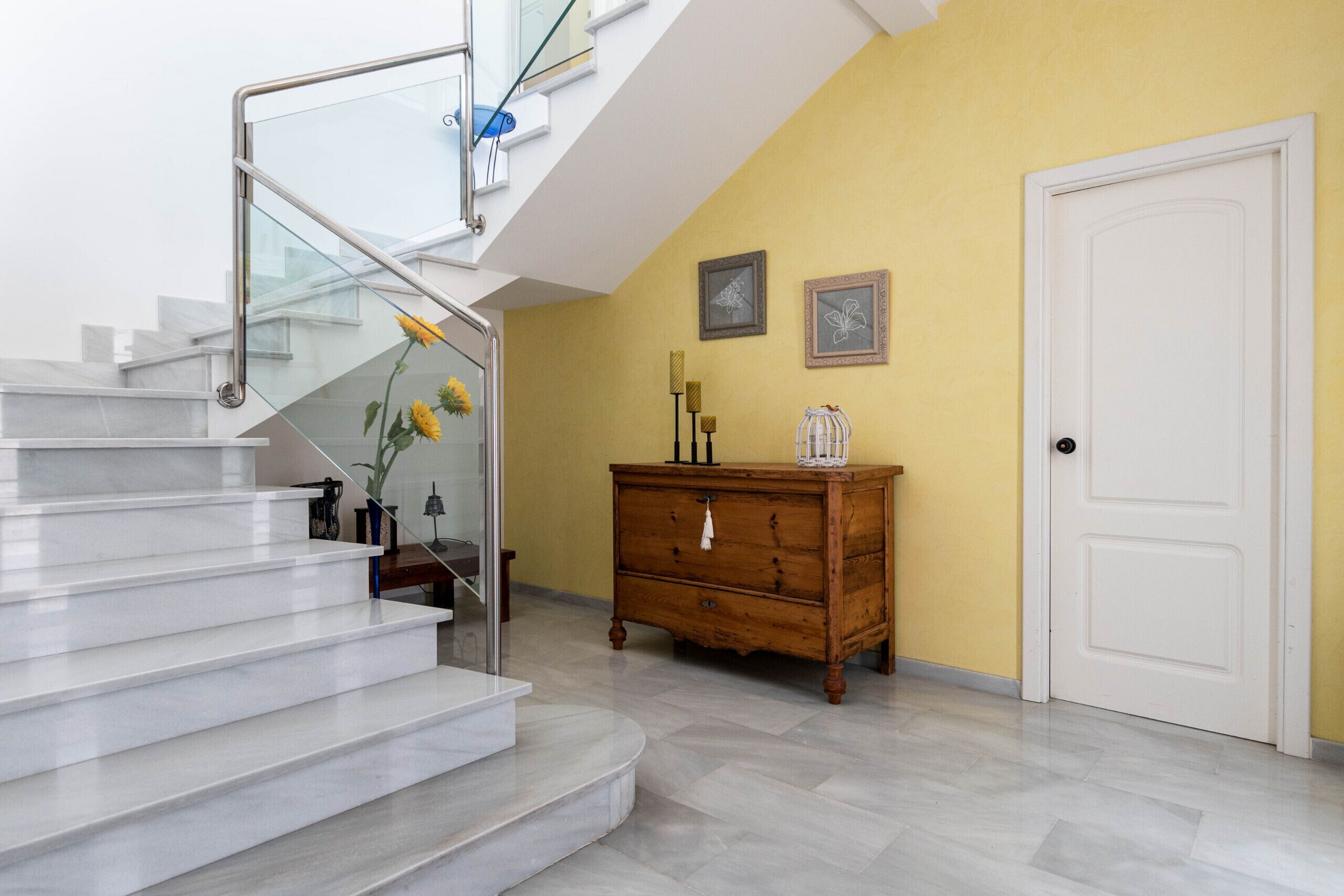 A staircase with glass railings leading to a second floor, a wooden dresser, and a white door in a room with yellow walls.