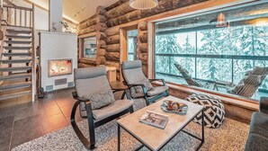 A log cabin interior with a fireplace, two armchairs, a coffee table, and a large window offering a snowy mountain view. - Villa rytilampi b by Interhome (Kuusamo)