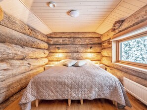 A log cabin bedroom with a bed, pillows, a nightstand, a window with a view of snow-covered trees, and a ceiling light. - Villa rytilampi b by Interhome (Kuusamo)