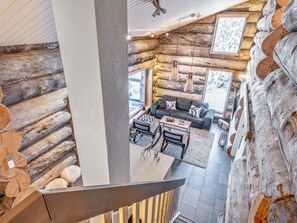 Interior de uma cabana de toras com escada, sala de estar com sofá e mesa de centro, e vista para o exterior.