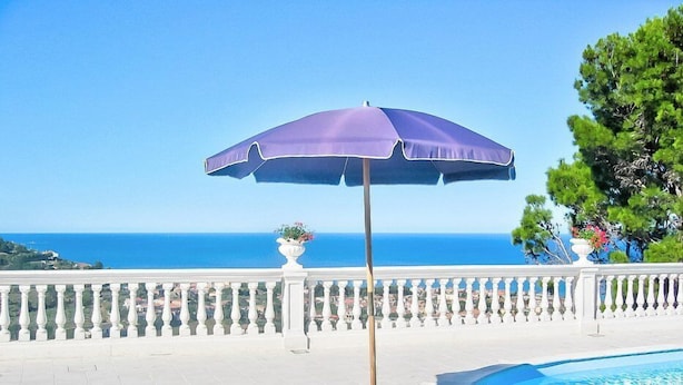 Zona de piscina con vista al mar, sillas de descanso azules, una sombrilla morada y una mesa con bebidas y fruta.