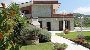 Una casa de dos pisos con fachada de piedra, un balcón y un jardín con plantas en macetas y flores.