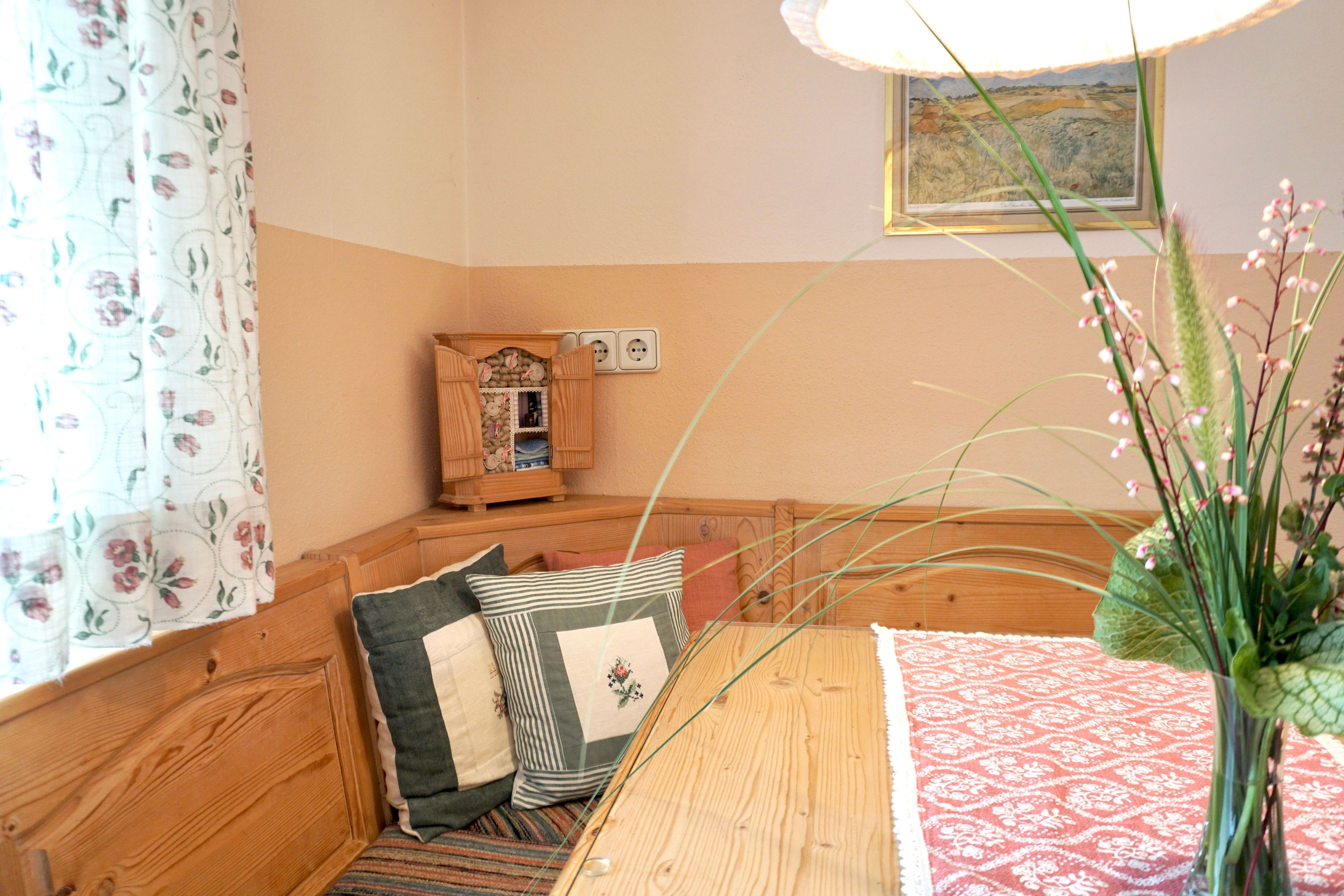 Eine Holzbank mit Kissen, ein kleiner Holzkasten, eine Vase mit Blumen und ein gerahmtes Bild an der Wand.