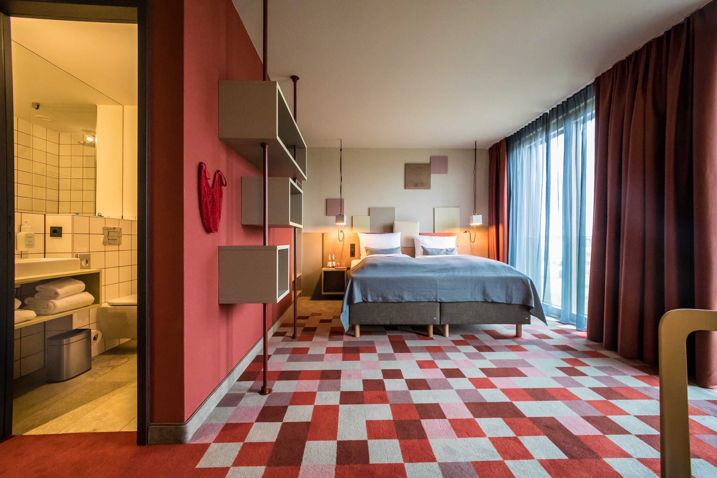 Une chambre d’hôtel avec un sol à carreaux, un lit, une table de chevet, un miroir, un mur rouge et une fenêtre avec des rideaux.