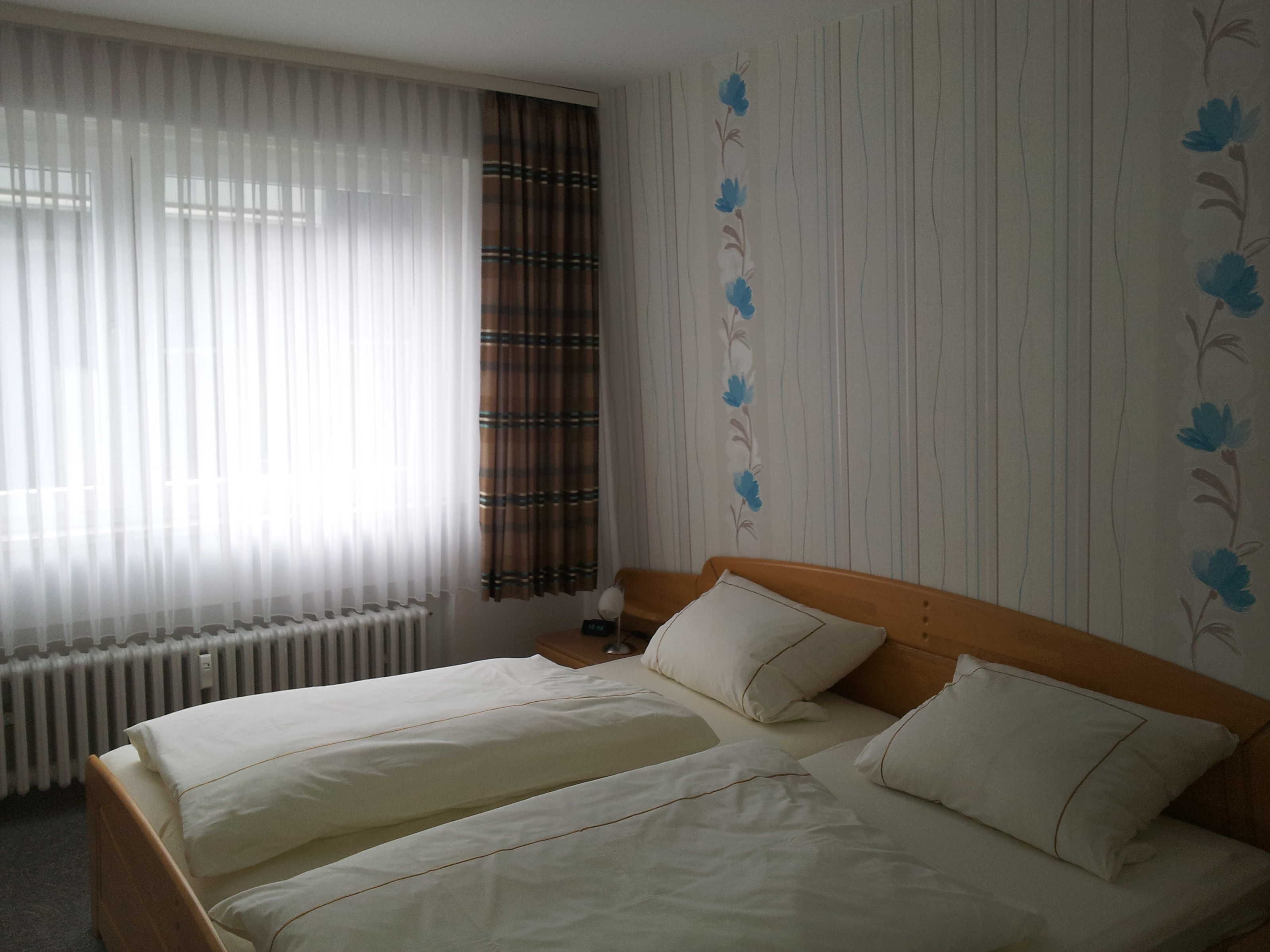 Ein Doppelbett mit weißen Bettwäsche in einem Zimmer mit Streifenmuster-Tapete und einem Fenster mit durchsichtigen Vorhängen.