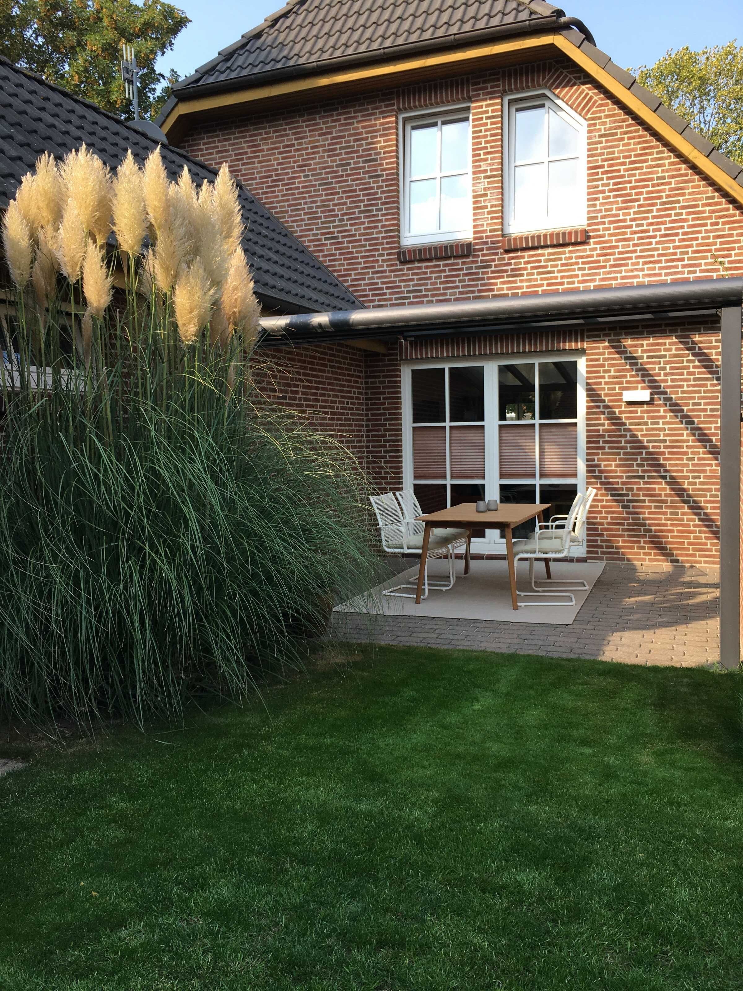 Een bakstenen huis met een overdekte patio, een tafel en stoelen, en hoge grassen.