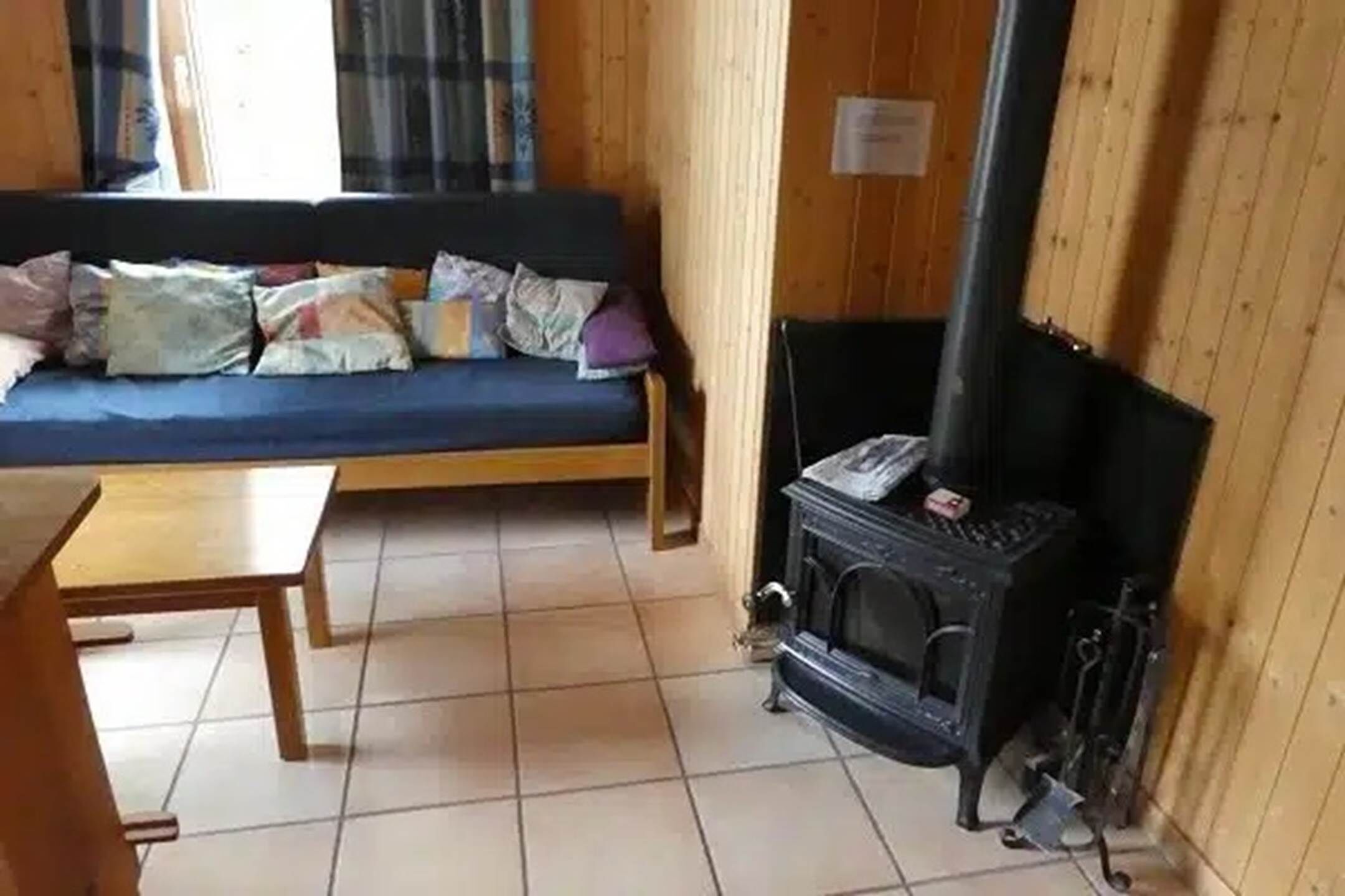 Una habitación con suelo de madera, un sofá con cojines, una mesita y un estufa de leña.