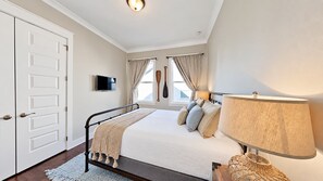 A bedroom with a bed, bedside lamp, curtains, a television, and a door. - The Den on 30A - 3BR/2.5BA Prominence Home - Sleeps 8 - Golf Cart & Ping Pong Table (Santa Rosa Beach)