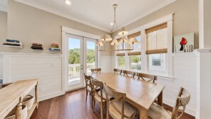 A dining area with a wooden table and chairs, a chandelier, and a view through the glass doors. - The Den on 30A - 3BR/2.5BA Prominence Home - Sleeps 8 - Golf Cart & Ping Pong Table (Santa Rosa Beach)