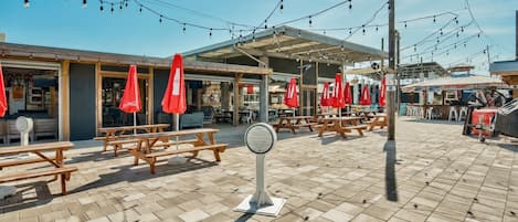 An outdoor seating area with picnic tables and red umbrellas, under a covered section with string lights.