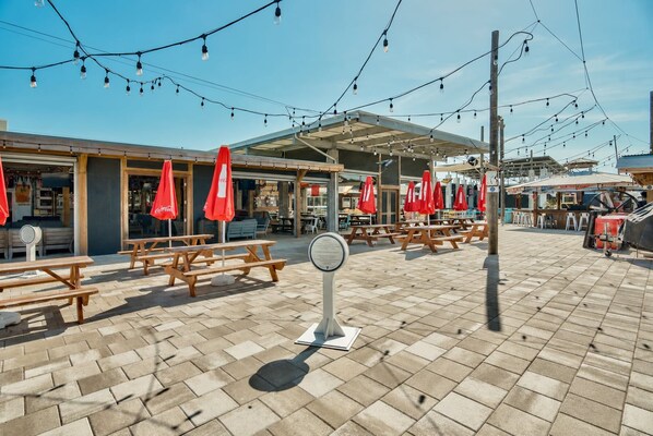An outdoor seating area with picnic tables and red umbrellas, under a covered section with string lights. - The Den on 30A - 3BR/2.5BA Prominence Home - Sleeps 8 - Golf Cart & Ping Pong Table (Santa Rosa Beach)