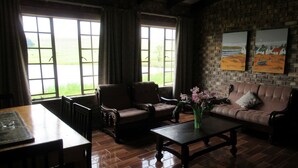Una habitación acogedora con pared de ladrillo, un sofá de cuero marrón, una mesa de madera y dos sillones. Amplias ventanas con cortinas ofrecen vistas a un paisaje verde.